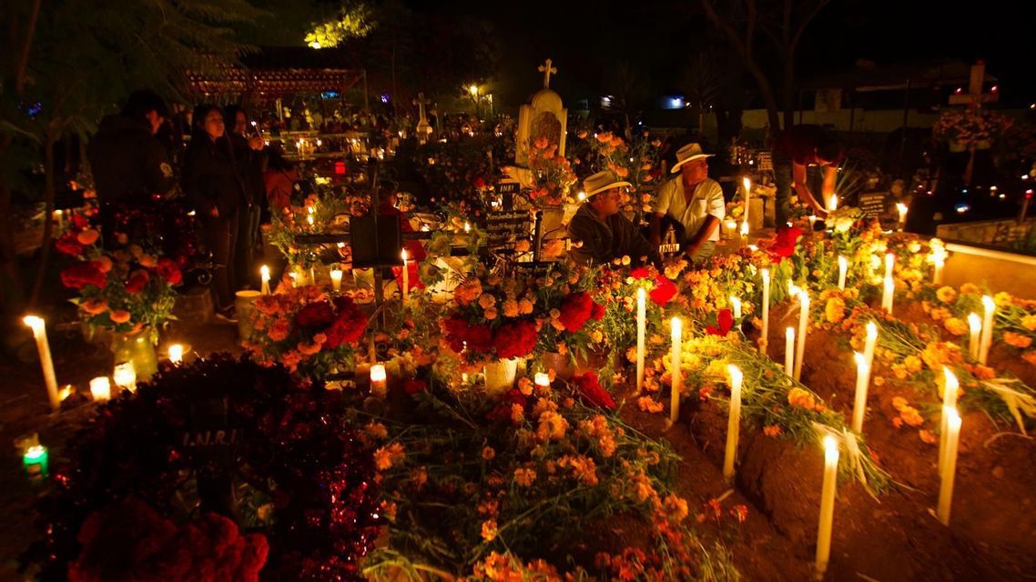 People gathered at night in a cemetery, where graves are covered with marigolds and lit by the warm glow of many tall candles.