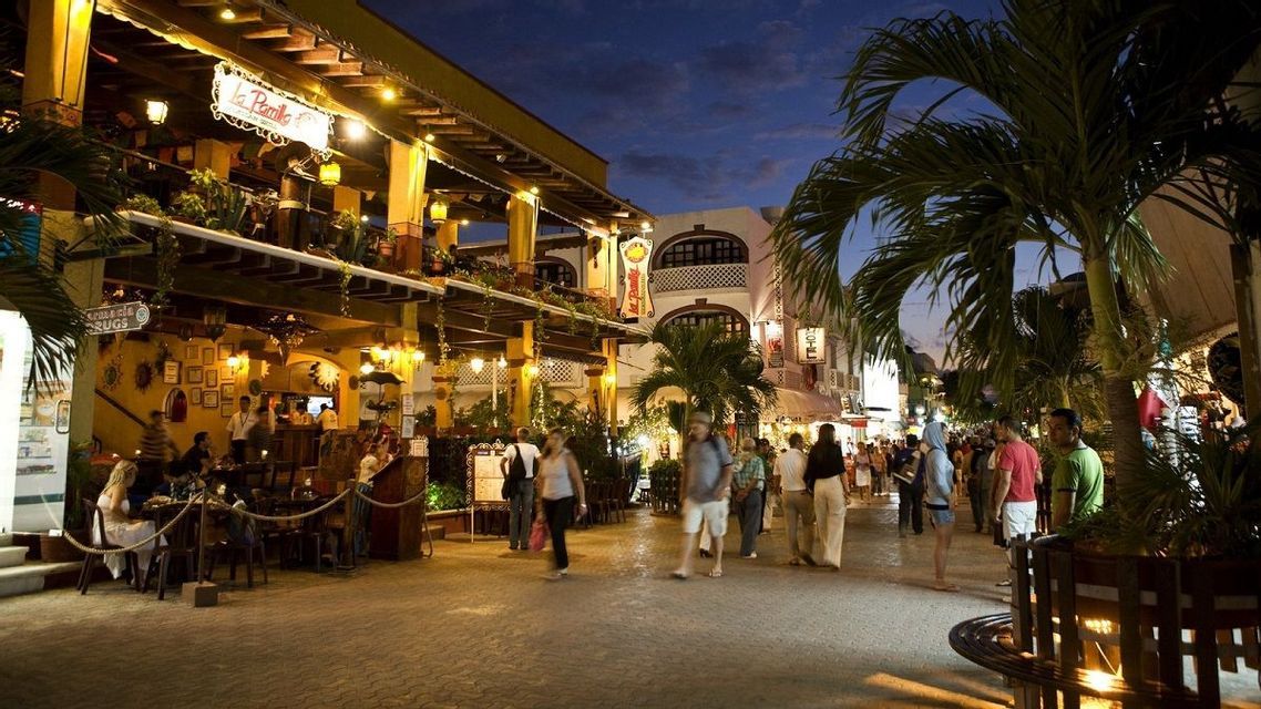 A bustling street scene at dusk, with people walking along a paved road lined with illuminated restaurants and palm trees.