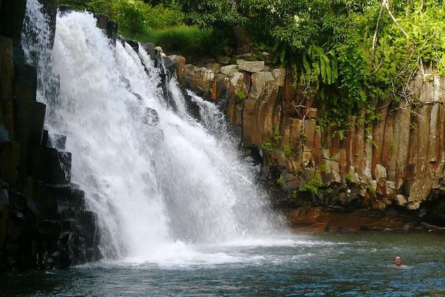 Eine Person schwimmt in einem Pool am Fuße eines breiten Wasserfalls, der über dunkle, säulenartige Felsen fließt und von grünem Laub umgeben ist.
