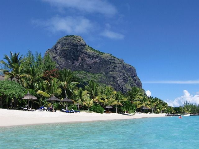 Ein tropischer weißer Sandstrand mit Palmen und Liegestühlen liegt an türkisfarbenem Wasser vor einem großen felsigen Berg.
