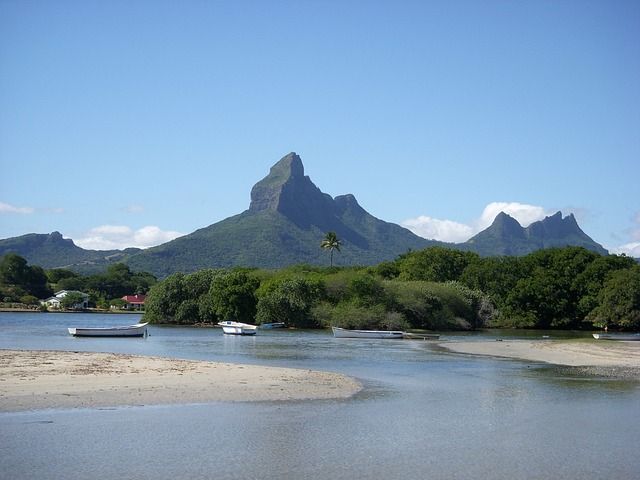 Boote liegen in einer ruhigen Bucht mit einer Sandbank vor einer üppigen, zerklüfteten Bergkette unter blauem Himmel vor Anker.