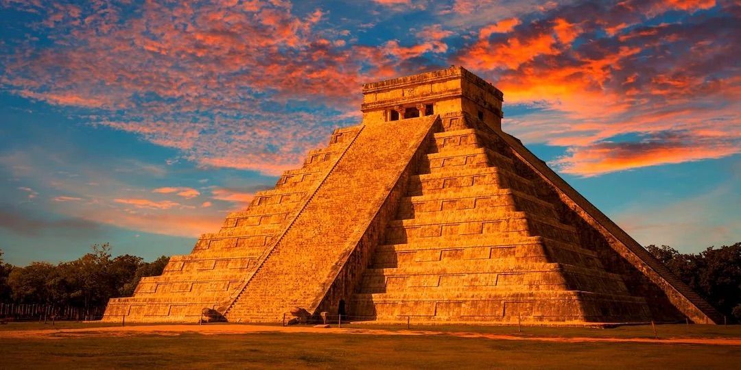 A large stone step-pyramid is illuminated by the setting sun, set against a blue sky with dramatic orange clouds.