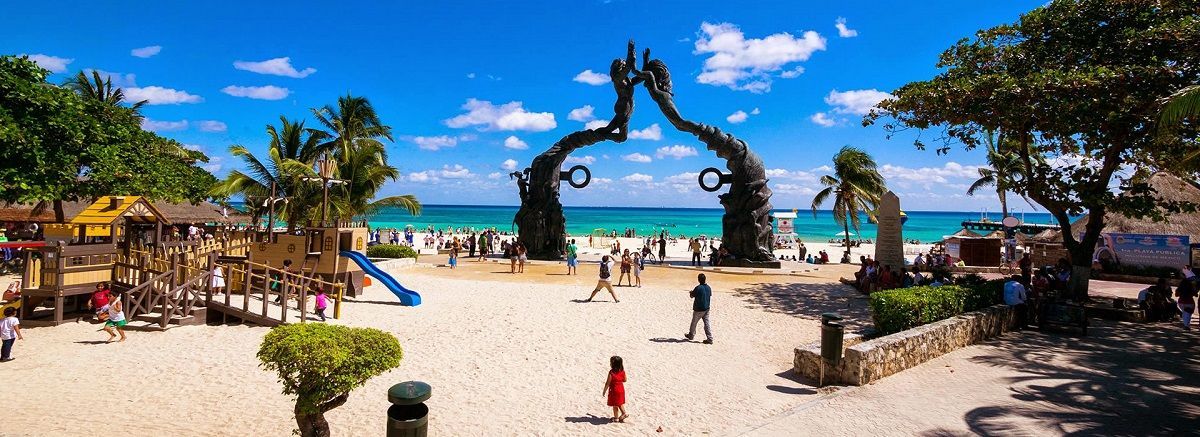 A large sculpture frames a busy tropical beach with people, a playground, and turquoise water under a blue sky.