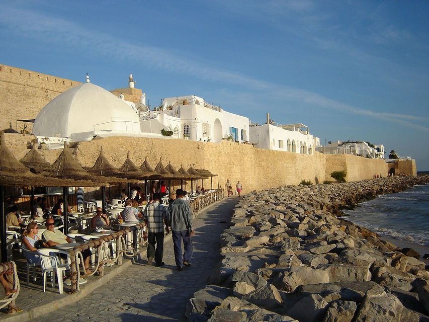 Des gens se promènent et s'installent à un café en plein air, avec des parasols de chaume, le long d'une promenade en pierre au bord de la mer, avec un mur fortifié et des bâtiments blancs en arrière-plan.