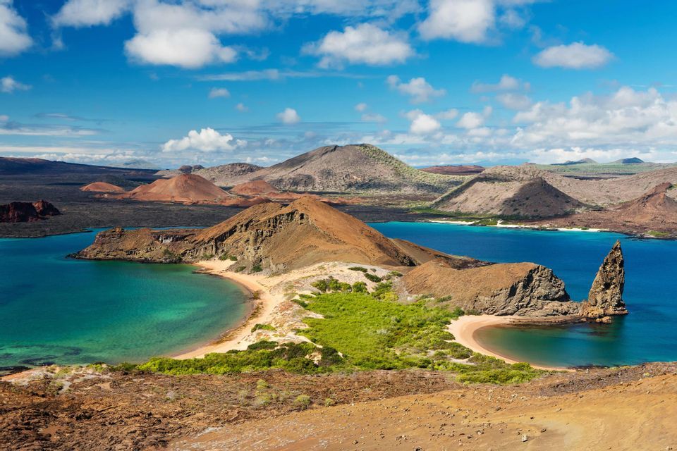 Una vista de islas volcánicas rodeando calas turquesas con playas de arena, un pináculo de roca y un cielo azul parcialmente nublado.