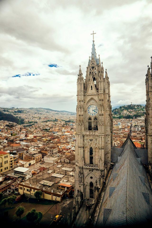 Un campanario gótico ornamentado de una catedral domina una extensa ciudad con colinas distantes bajo un cielo nublado.