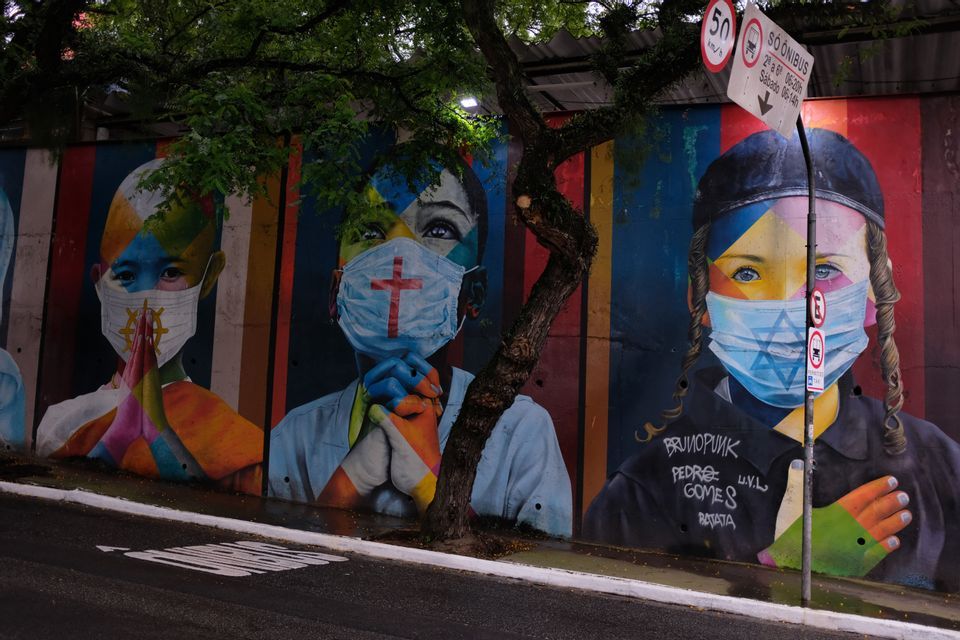 Ein farbenfrohes Street-Art-Wandgemälde an einer Stadtmauer, das drei Kinder mit Gesichtsmasken und verschiedenen religiösen Symbolen zeigt.