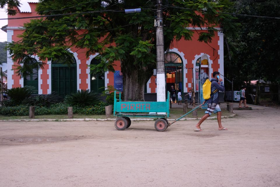 Un homme avec un sac à dos jaune tire une charrette verte le long d'une route sablonneuse devant un bâtiment colonial orange.