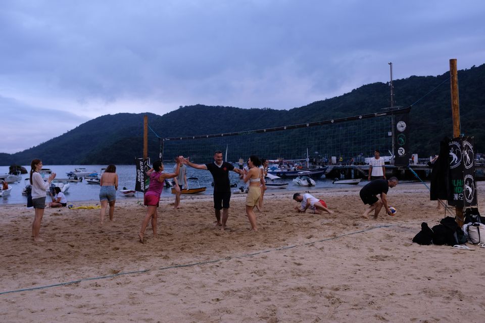 Un viaje en grupo de WeRoad juega voleibol de playa en una orilla arenosa con barcos en el agua y colinas al fondo.
