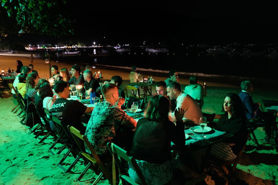Eine WeRoad-Gruppenreise beim Abendessen an langen Tischen an einem Sandstrand bei Nacht, Boote auf dem Wasser sichtbar.