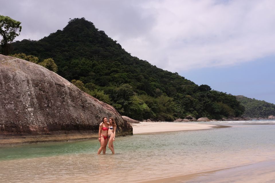 Zwei Frauen in Bikinis einer WeRoad-Gruppenreise stehen lächelnd im seichten Wasser am Strand, mit einem großen Felsen und bewaldetem Hügel im Hintergrund.