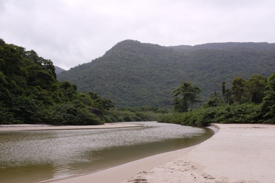 Ein Fluss schlängelt sich entlang eines sandigen Ufers mit dicht bewaldeten Hügeln im Hintergrund unter einem bewölkten Himmel.