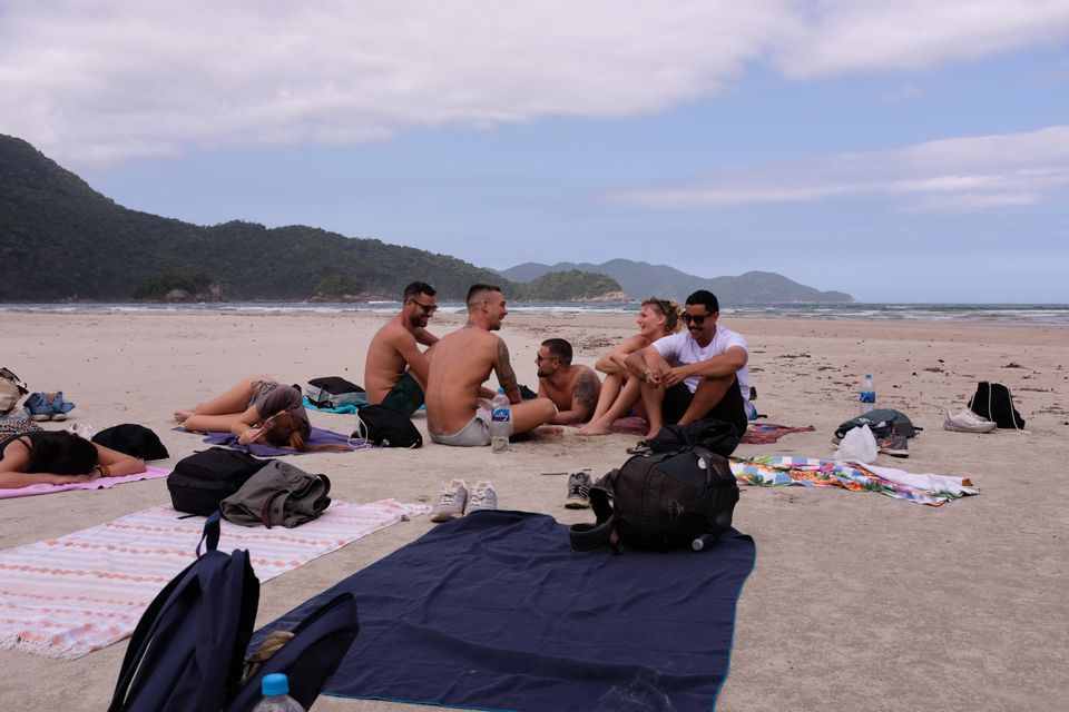 Eine WeRoad-Gruppe entspannt auf Handtüchern an einem breiten Sandstrand, mit grünen, bewaldeten Hügeln im Hintergrund auf der anderen Wasserseite.