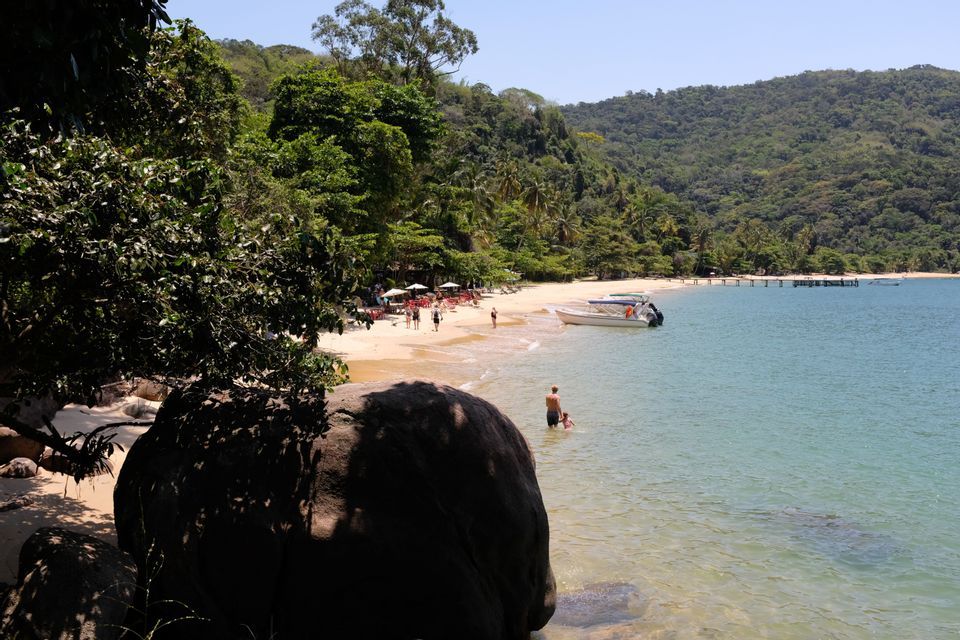 Menschen entspannen sich an einem Sandstrand neben türkisfarbenem Wasser, mit einem vertäuten Boot und einem üppigen, bewaldeten Hügel im Hintergrund.
