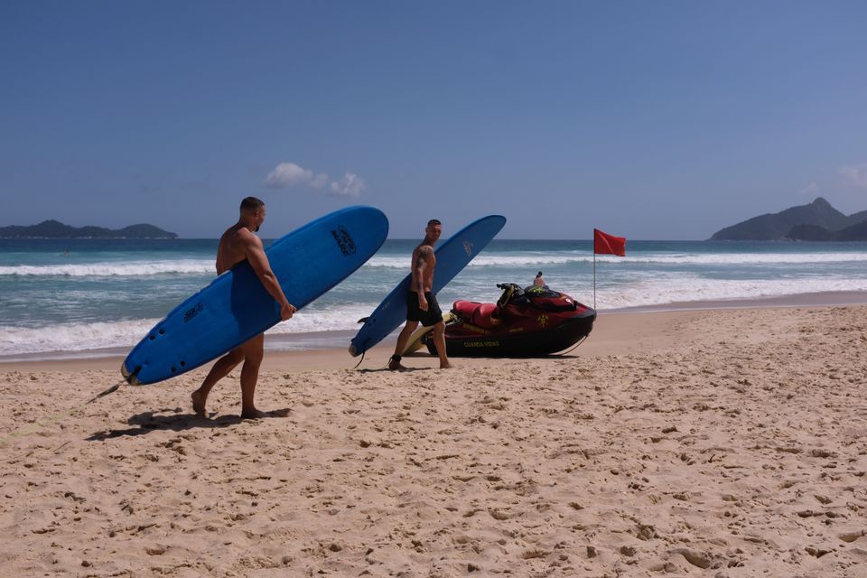 Zwei Männer tragen blaue Surfbretter, während sie an einem Sandstrand neben dem Ozean entlanggehen, mit einem Jetski in der Nähe geparkt.