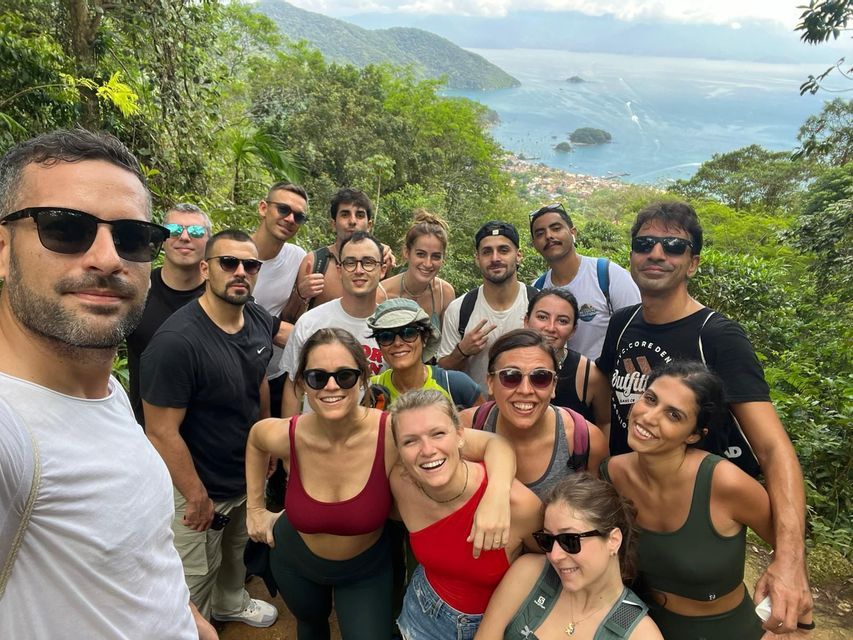 Un grupo de viaje de WeRoad posa para una selfie en una exuberante ladera verde con vistas a una bahía costera y montañas.