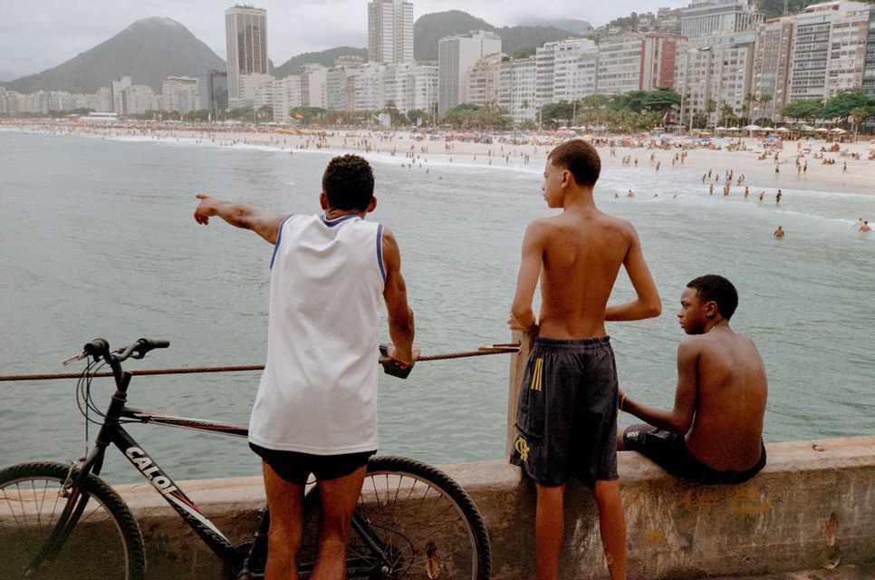Drei junge Männer stehen mit einem Fahrrad von hinten auf einem Steg und blicken auf einen belebten Strand und eine Stadtsilhouette.