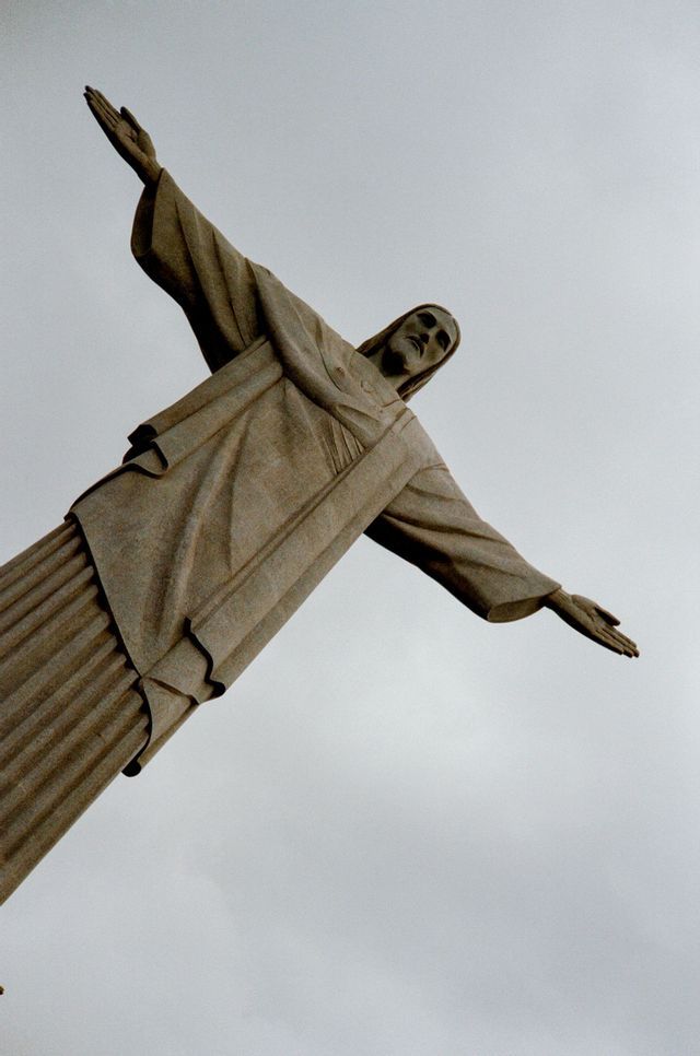 Eine diagonale Untersicht der Christus-Erlöser-Statue mit ausgebreiteten Armen vor einem bedeckten Himmel.