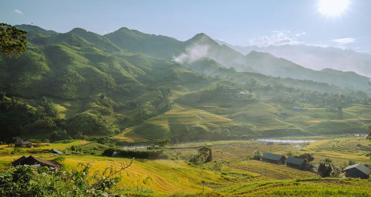 Una vasta valle con campi di riso terrazzati su dolci colline verdi, case e montagne nebbiose sotto un sole splendente.