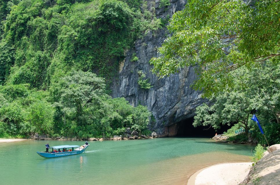 Una barca con persone a bordo naviga su un fiume turchese verso l'ampio ingresso di una grotta, alla base di una scogliera ricoperta dalla giungla.