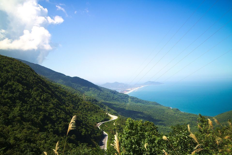 Vista panoramica di una strada tortuosa su una montagna verdeggiante che domina una costa sabbiosa e il mare blu.