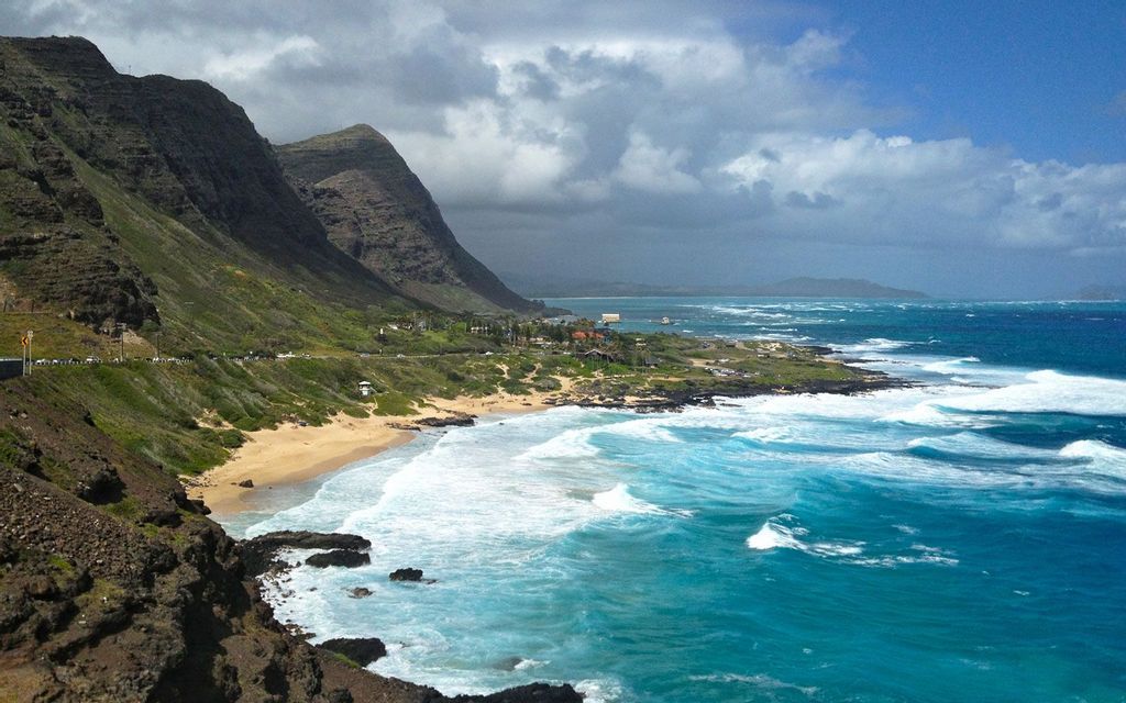 Una costa verde e frastagliata con una spiaggia sabbiosa incontra l'oceano turchese con onde bianche sotto un cielo nuvoloso.