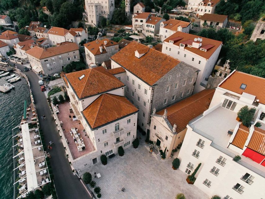 Veduta aerea di una città costiera con edifici in pietra, tetti rossi e una piazza sul lungomare con ristoranti all'aperto.
