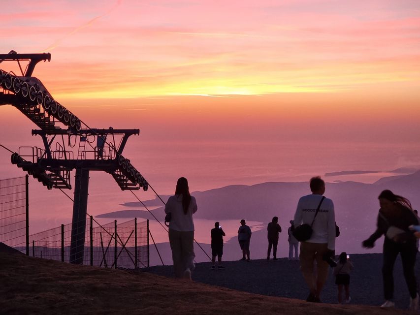 Un viaggio di gruppo WeRoad per ammirare il tramonto da una cima di montagna, accanto a un impianto di risalita, stagliati contro un cielo rosa e arancione.
