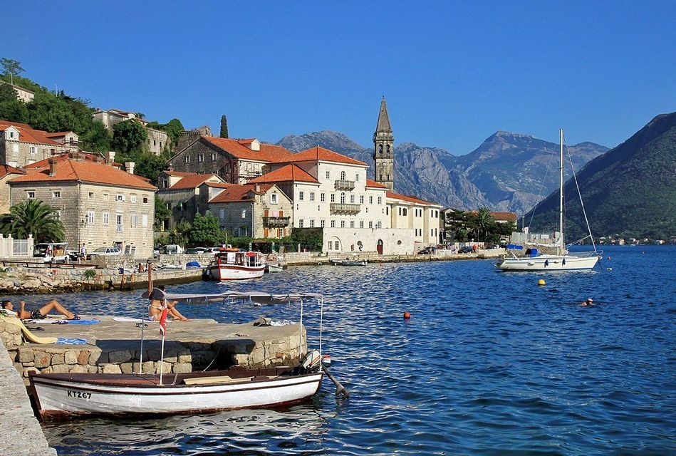 Una città costiera con edifici in pietra e un campanile si affaccia su una baia blu, con barche sull'acqua e montagne sullo sfondo.