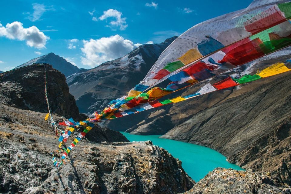 Bandiere di preghiera colorate che sventolano al vento su un fiume turchese in una ripida valle di montagna rocciosa sotto un cielo blu.