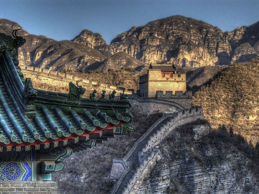 Una torre di guardia sulla Grande Muraglia che serpeggia su una montagna rocciosa, con un dettagliato tetto di tempio con tegole verdi in primo piano.