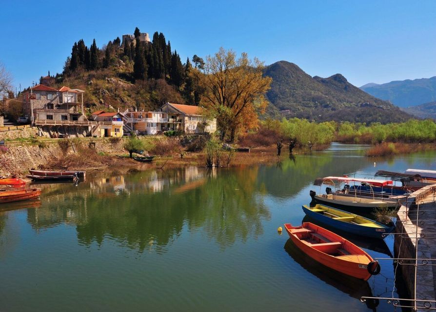 Barche colorate ormeggiate su un lago calmo di fronte a un villaggio collinare con una fortezza sulla cima sotto un cielo sereno.