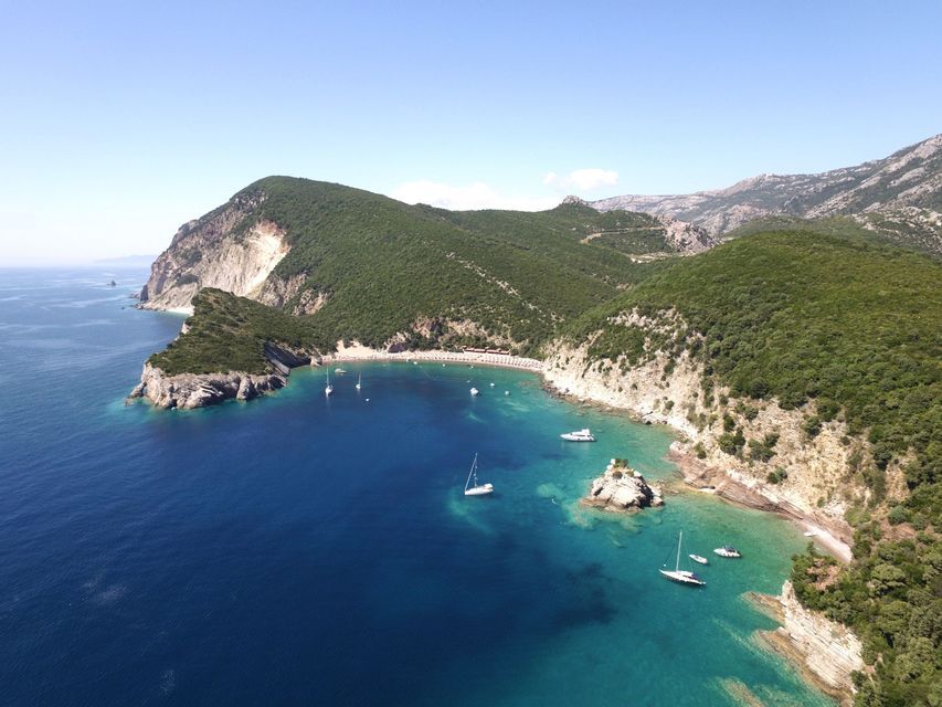 Vista aerea di una baia turchese con barche a vela all'ancora, racchiusa tra ripide scogliere alberate sotto un cielo azzurro e limpido.