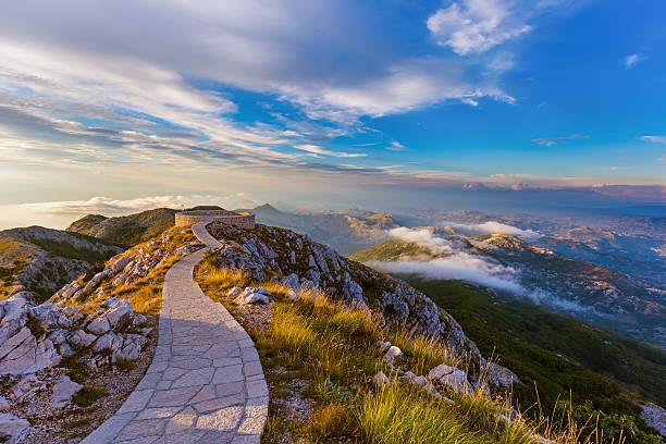 Un sentiero lastricato in pietra si snoda lungo un crinale roccioso di montagna verso un punto panoramico circolare, che si affaccia su una vasta valle con nuvole al di sotto.