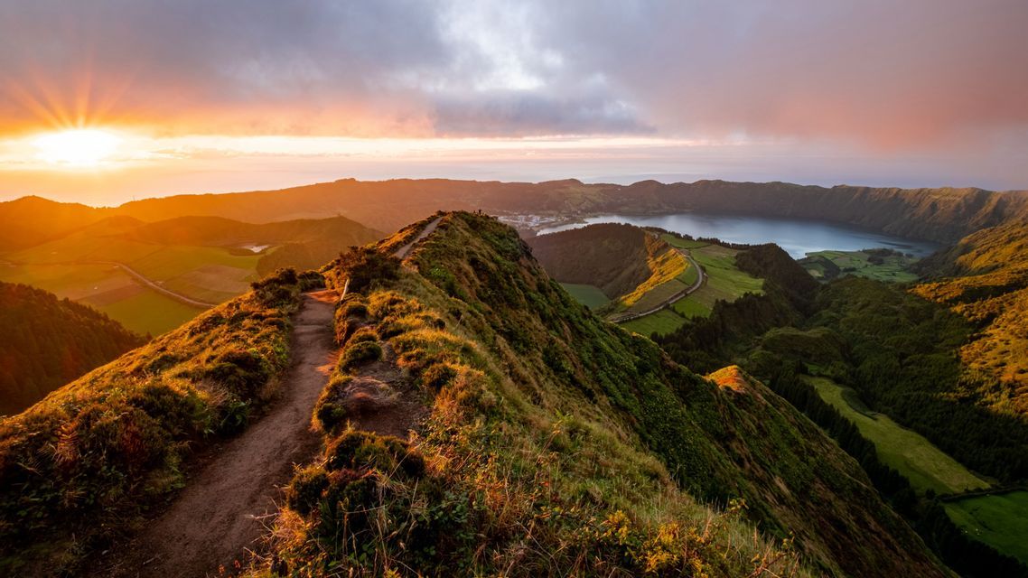 Un sentier de randonnée le long d'une crête montagneuse herbeuse surplombe un lac de cratère et l'océan au coucher du soleil doré.