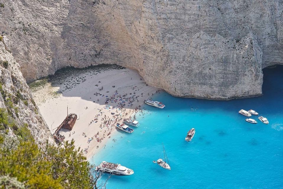 Una vista aérea de un naufragio en una cala de playa de arena, con barcos en el mar turquesa y gente tomando el sol en la orilla.