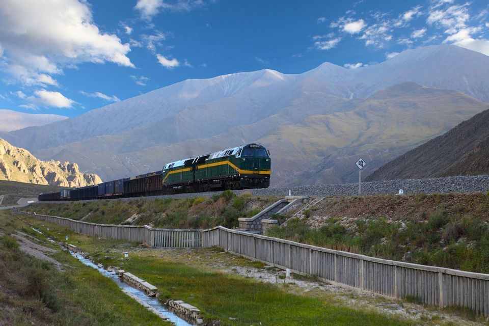 Un treno merci verde e giallo viaggia su un binario attraverso una vasta valle di montagna sotto un cielo blu con nuvole bianche.