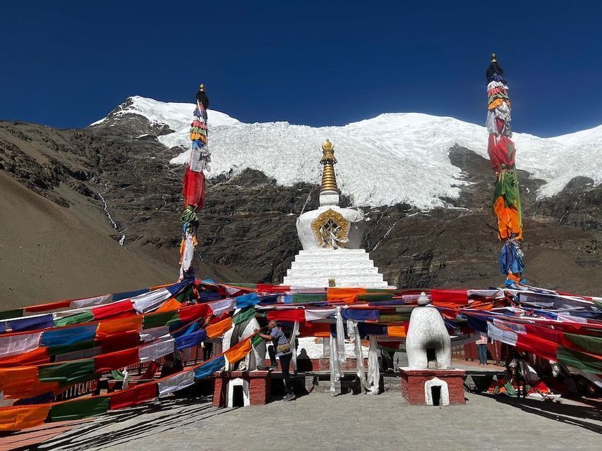 Un bianco stupa buddista è adornato con bandiere di preghiera colorate, con una grande montagna innevata sullo sfondo sotto un cielo sereno.