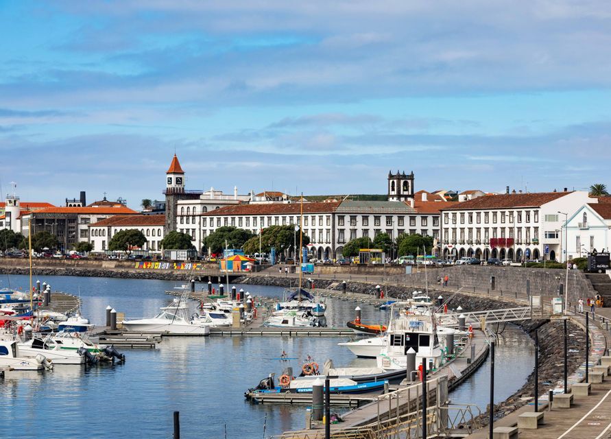 Une marina remplie de bateaux amarrés le long d'un quai en pierre, avec une ville côtière de bâtiments blancs et une tour de l'horloge en arrière-plan.