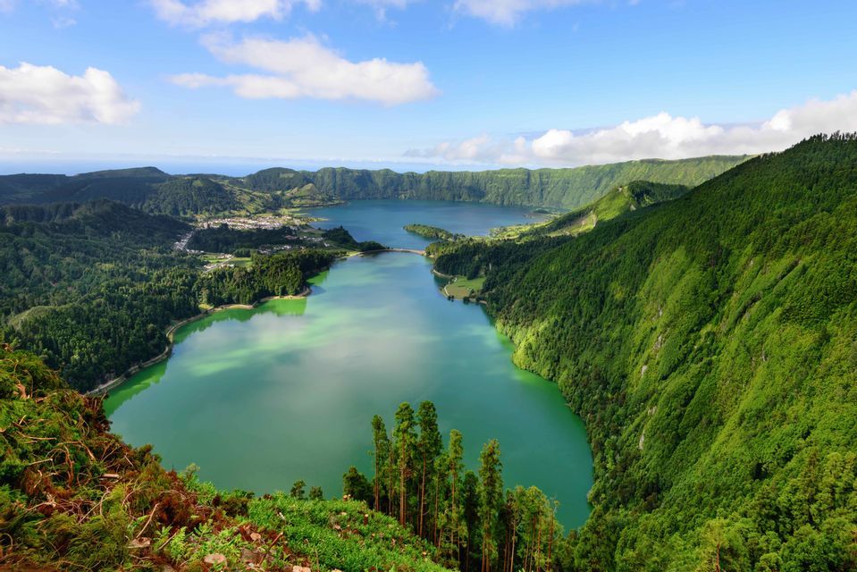 Vue aérienne de deux grands lacs de cratère connectés, entourés de pentes verdoyantes et escarpées, sous un ciel bleu nuageux.