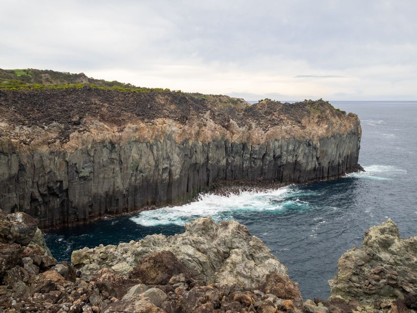 Une haute falaise de basalte en colonnes rencontre la mer sombre, avec des vagues blanches s'écrasant contre sa base rocheuse.