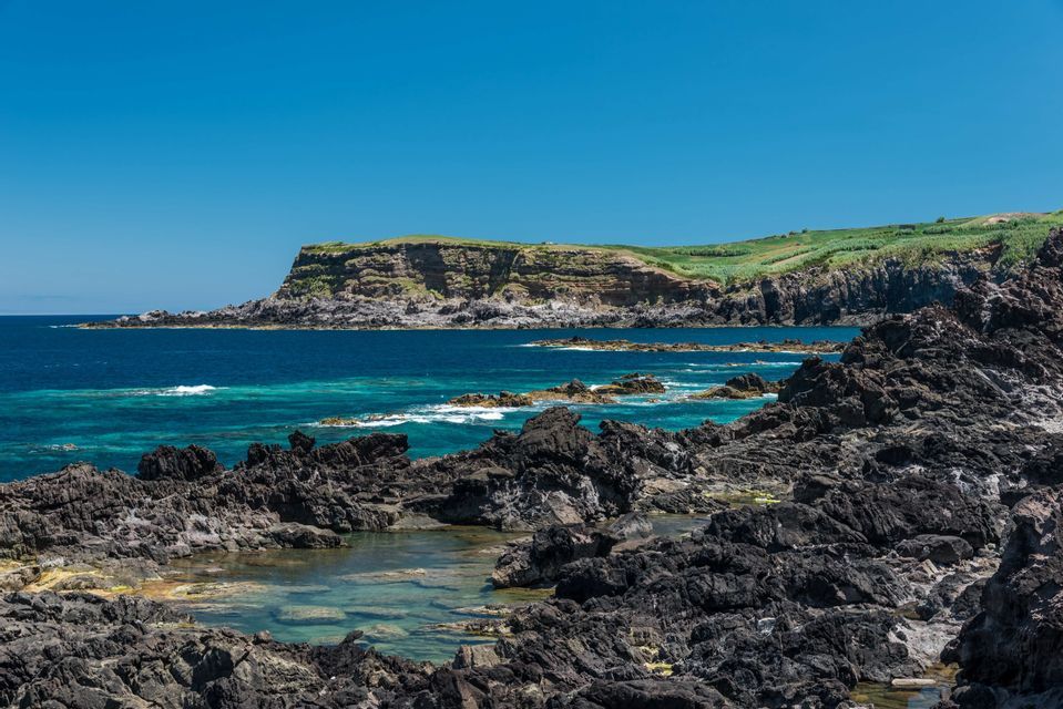 Un littoral rocheux et volcanique avec des piscines naturelles, une mer turquoise et des falaises vertes lointaines sous un ciel bleu clair.