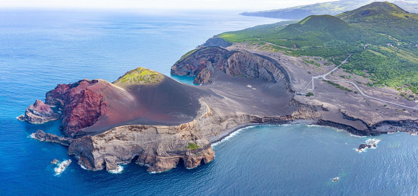 Vue aérienne d'une péninsule volcanique avec terre rouge et noire, rencontrant des falaises escarpées et l'océan d'un bleu profond.