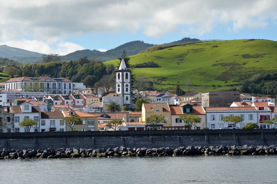 Une ville côtière avec des bâtiments colorés et un clocher d'église sombre se dresse derrière une digue en pierre, avec une grande colline verte en arrière-plan.