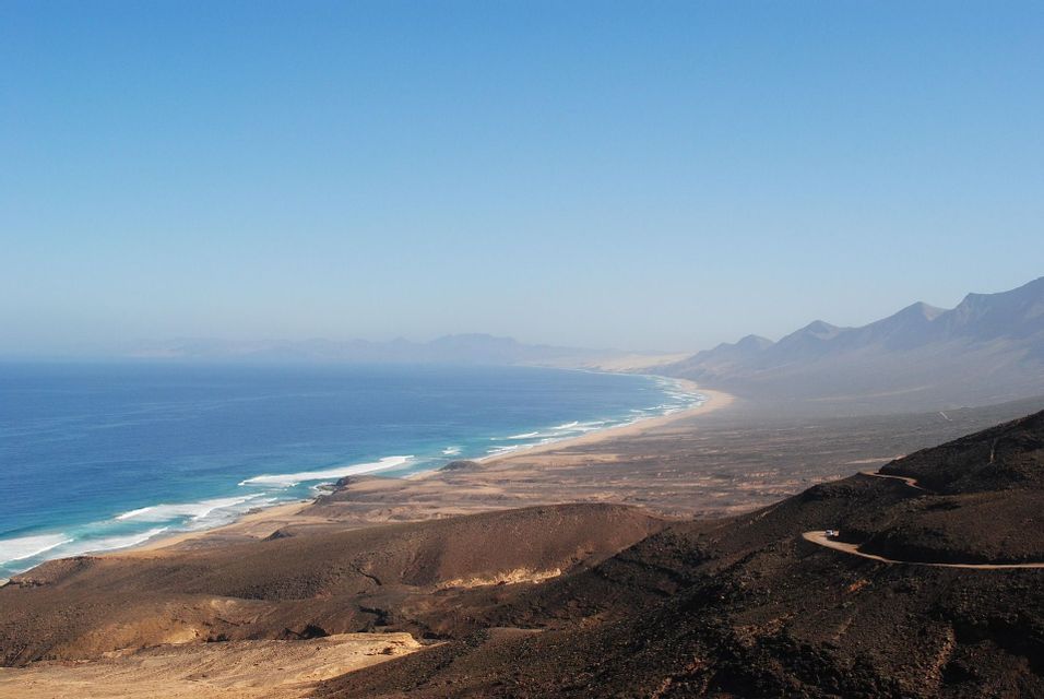 Una extensa costa vista desde arriba, con una playa de arena, olas azules del océano y una carretera sinuosa a través de montañas áridas bajo un cielo despejado.