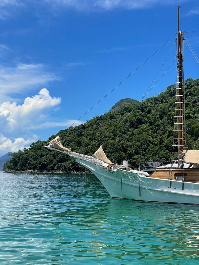 La prua di una barca a vela bianca galleggia su acque tranquille e turchesi vicino a un'isola lussureggiante e alberata sotto un cielo azzurro.