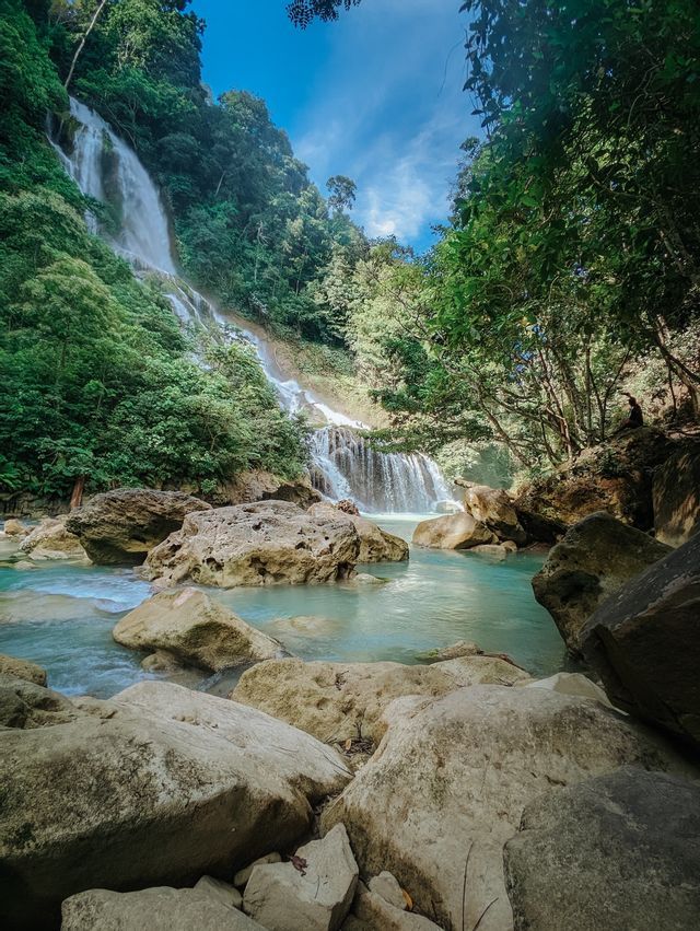 Un'alta cascata scende da un fianco boscoso in una pozza rocciosa dalle acque turchesi, con una persona seduta su una roccia lì vicino.