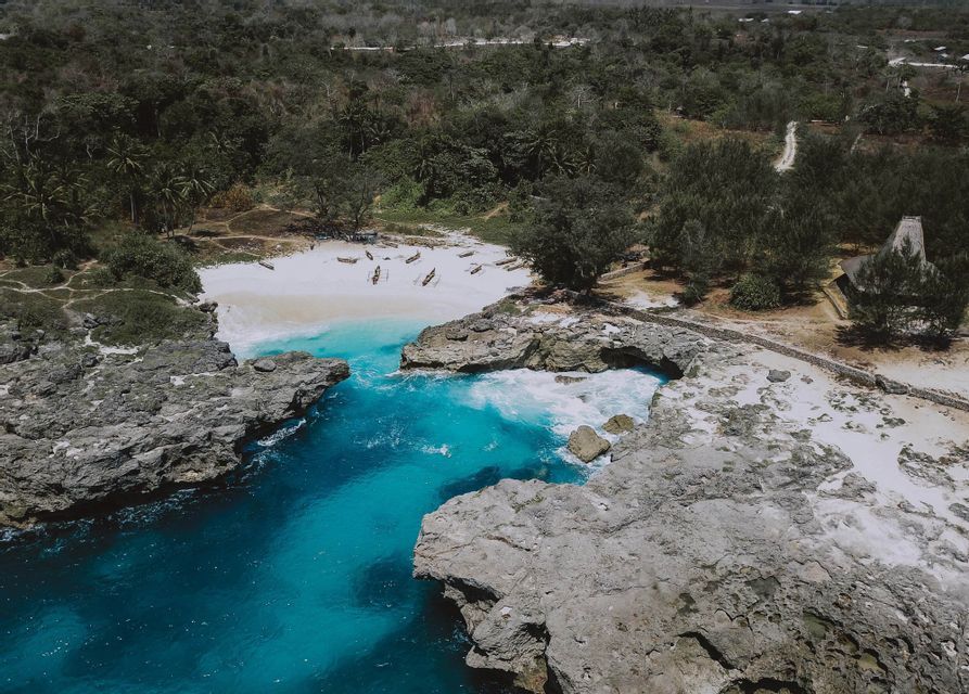 Una vista aerea di una laguna turchese e una spiaggia di sabbia bianca con barche, delimitata da scogliere rocciose e una fitta foresta.
