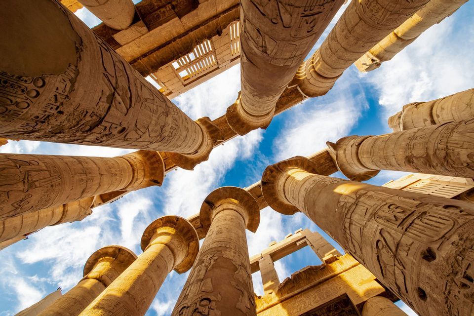 Una vista en contrapicado de enormes columnas antiguas con jeroglíficos que se elevan hacia un cielo azul con nubes.
