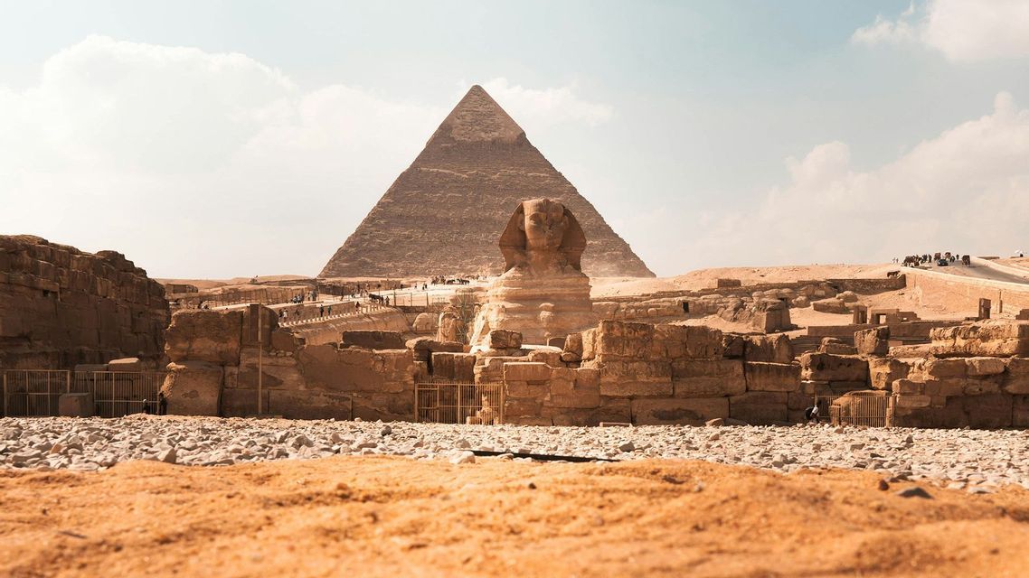 Una vista desde el suelo de la Gran Esfinge con una gran pirámide al fondo, rodeada de antiguas ruinas de piedra bajo un cielo brillante.